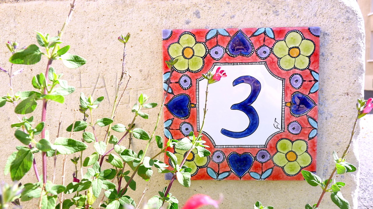 Numéro de maison céramique rouge avec motifs cœur fleurs bleus vertes violettes sur mur pierre blanc, jolies plantes roses design personnalisable original